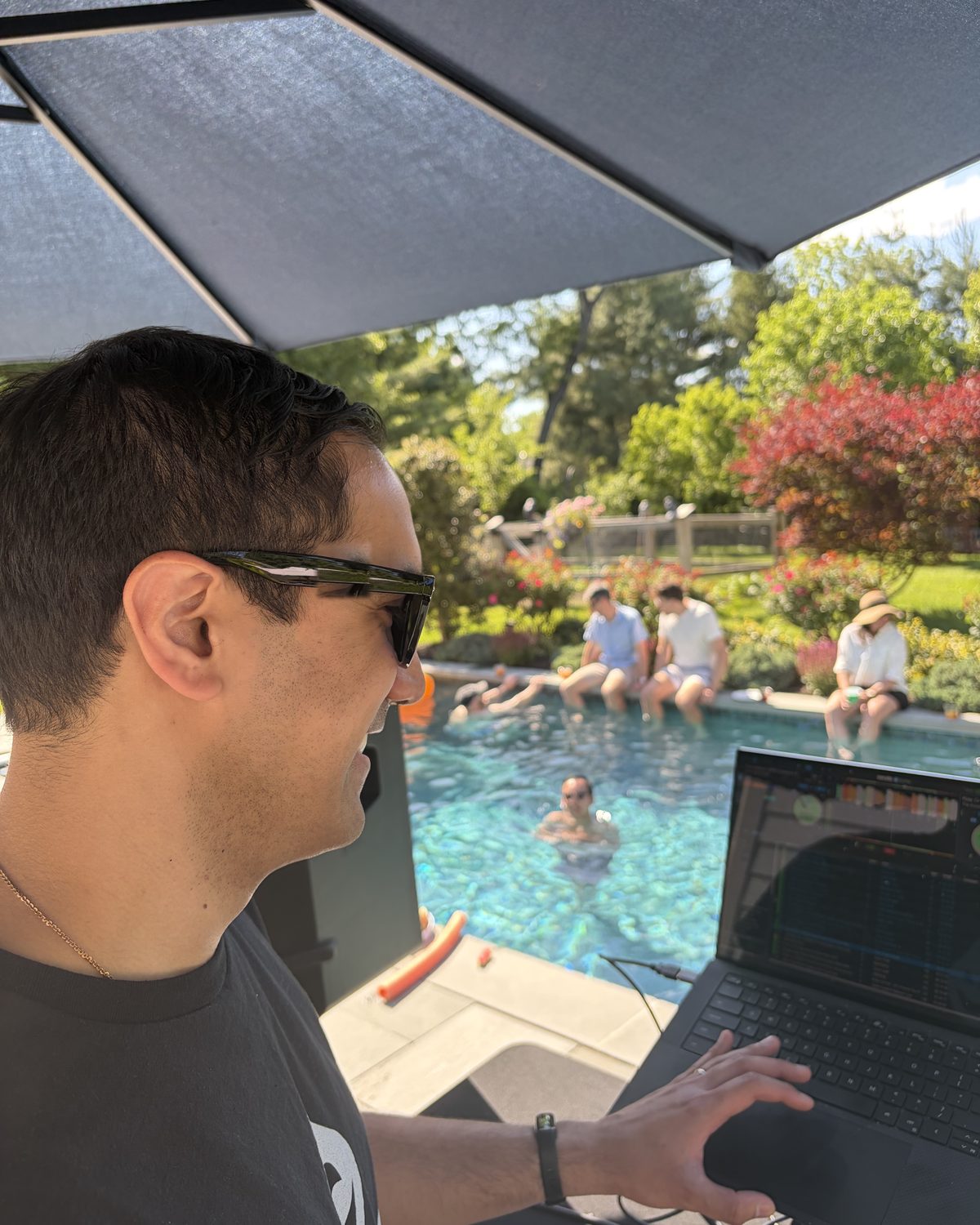 DJ idrop in profile behind a laptop at a pool party, guests in the water behind him
